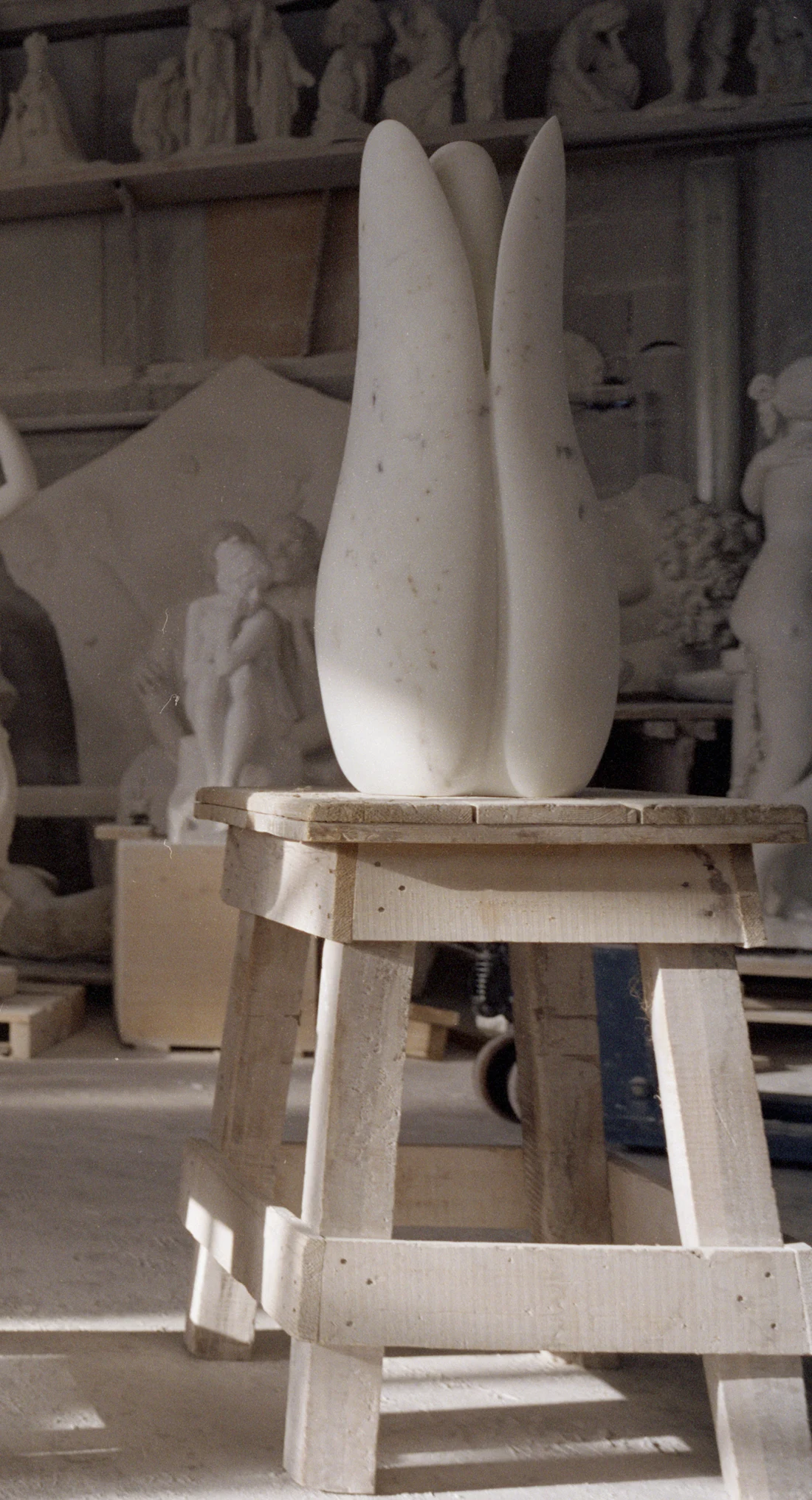 A small white vertical marble sculpture sitting on a rough wooden stool in a dusty atelier environment
