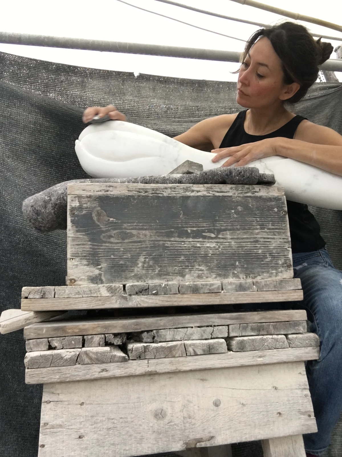 The sculptor Yoko Kubrick sanding and finishing a white marble sculpture, leaning over weathered wooden blocks in her studio